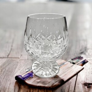 May include: A clear crystal glass with a cut design sits on a wooden coaster. The glass is empty and appears to be for serving liquor.