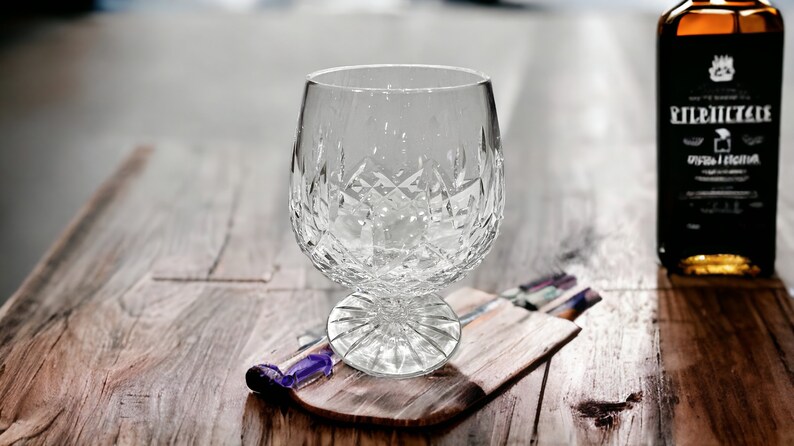 May include: A clear crystal glass with a cut design sits on a wooden coaster. The glass is empty and appears to be for serving liquor.