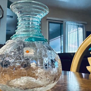 Può includere: Un vaso di vetro trasparente fatto a mano con un motivo craquelé e un bordo turchese. Il vaso ha una base bulbosa e un collo strutturato. L'etichetta sul vaso recita "HANDMADE". Il vaso è appoggiato su un tavolo di legno.