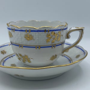 May include: A white porcelain teacup and saucer set with a blue and gold floral pattern. The cup has a scalloped rim and a delicate handle.