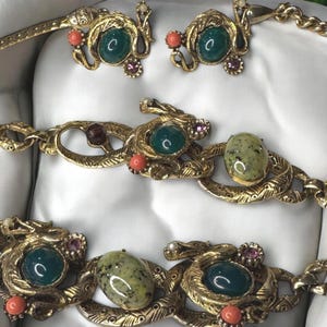 May include: A gold-tone snake-shaped necklace and earrings set with green, coral, and purple stones. The necklace features a repeating pattern of snake heads with stones set in the eyes.