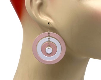 May include: A pair of pink and white circle earrings with a gold chain hanging from the top. The earrings are layered with three circles, the largest being pink, the middle being white, and the smallest being pink.