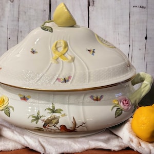 Puede incluir: Una fuente de servir de porcelana blanca con tapa, decorada con un motivo de limón, mariposas y diseños florales. La fuente tiene dos asas y un borde dorado. La tapa presenta una cinta amarilla y un limón en la parte superior. La fuente está rodeada de limones sobre un paño blanco.