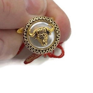 May include: Gold ring with a white pearl center featuring a gold bull head design. The ring is surrounded by a decorative gold border.