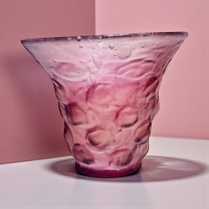 Può includere: Un vaso di vetro rosa con un motivo strutturato a bolle. Il vaso ha un'ampia apertura svasata e una base rosa più scura. Lo sfondo è di colore rosa pieno, creando un'estetica monocromatica. Il vaso è probabilmente utilizzato per scopi decorativi.