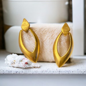 May include: A pair of gold-tone earrings with a teardrop shape and a cutout design. The earrings are on a white surface.