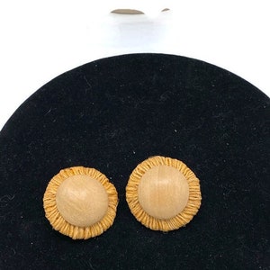 Può includere: Due orecchini rotondi marrone chiaro con un centro in legno e un bordo strutturato intrecciato. Gli orecchini sono esposti su una superficie nera su sfondo bianco.