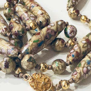 May include: Close-up of a vintage necklace featuring cream-colored, elongated beads with floral designs in pink, green, and blue, accented with gold-colored metal. The necklace includes round beads and a decorative gold-colored clasp.