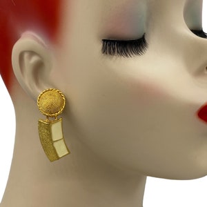 Könnte beinhalten: Goldfarbene Ohrhänger mit einem weißen Emaille-Detail. Die Ohrringe haben eine runde Oberseite und eine rechteckige Unterseite.