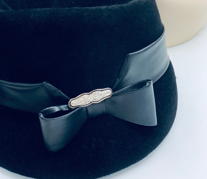 May include: A black felt hat with a black leather band and a silver bow. The bow has a silver pin with a decorative design.