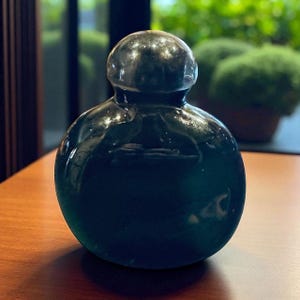 May include: A dark green, spherical glass bottle with a rounded top. The bottle has a glossy finish and sits on a wooden surface. The background is blurred, with hints of greenery visible through a window.