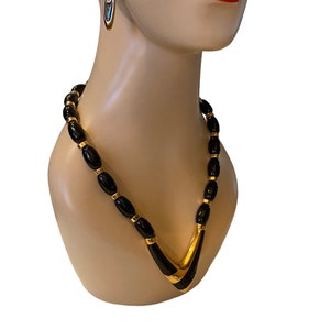May include: A gold and black beaded necklace with a V-shaped pendant. The necklace is made of black beads with gold accents. The pendant is made of gold and has a black enamel inlay.
