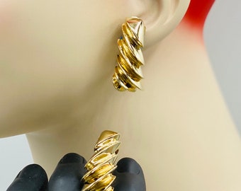 May include: A pair of gold-toned earrings with a spiral design. The earrings are shown on a mannequin's ear and hand.