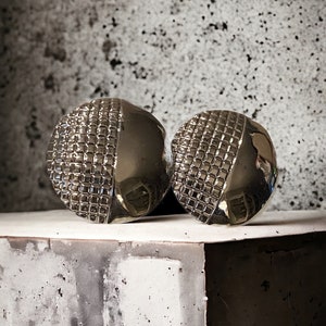 May include: A pair of silver tone earrings with a geometric design. The earrings are round and have a textured surface.