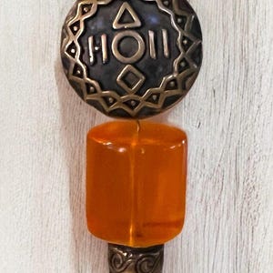 May include: A round, brass-colored pendant with a geometric design and the word "HOII" inscribed. The pendant is attached to a long, orange glass bead.