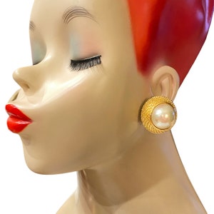 Può includere: Un paio di orecchini a clip dorati con un centro di perla finta. Gli orecchini sono rotondi e hanno un design testurizzato.