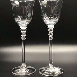 Könnte beinhalten: Zwei klare Glas-Stielgläser mit einem dekorativen, facettierten Stiel. Die Gläser stehen auf einer schwarzen Oberfläche. Der Boden eines Glases trägt den Schriftzug "Mikasa".