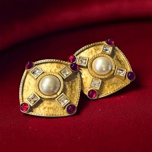 May include: A pair of gold-toned square earrings with a central pearl and surrounding red and clear gemstones. The earrings have a textured surface and are set against a red background.