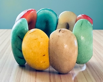 Pode incluir: Uma pulseira colorida com contas em forma de fruta. As contas são em tons de verde, amarelo, vermelho e bege. A pulseira é exibida em uma superfície de madeira marrom claro.