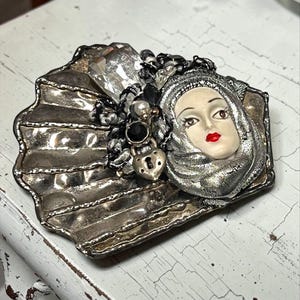 May include: A decorative brooch shaped like a silver-toned shell, featuring a central sculpted face with red lips, framed by a silver head covering. The piece is adorned with a large clear crystal, black beads, a small white pearl, and a heart-shaped charm.