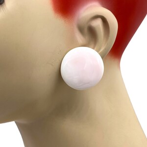 May include: A pair of white, round, clip-on earrings. The earrings are made of a smooth, opaque material and have a slightly pink hue.