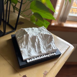 May include: A white 3D-printed mountain model, labeled "Schönteldspitze" in white text, sits on a black base. The model is highly detailed, showing the peaks and valleys of the mountain range. The base is on a yellow table.