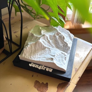 以下が含まれることがあります： ユングフラウ山脈の3Dプリントモデル。モデルは白で、黒いベースの上に「Jungfrau」という名前が白い文字で書かれています。モデルは詳細で、山の峰と谷を示しています。