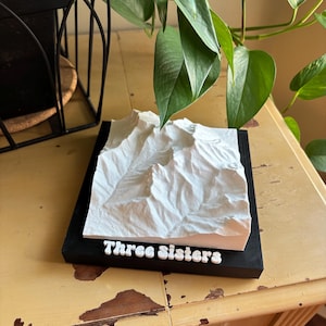 May include: A 3D-printed model of a mountain range, with the text "Three Sisters" in white letters. The model is white and sits on a black rectangular base. The model is displayed on a yellow table with a green plant in the background.
