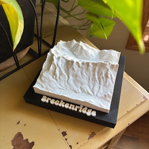 May include: A white 3D topographical model of a mountain range, mounted on a black base. The word "Breckenridge" is spelled out in white letters on the base. The model is likely a decorative item or souvenir.