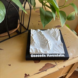 May include: A white 3D-printed model of a mountain range, labeled "Cascade Mountain," sits on a black base. The model is detailed with textured peaks and valleys. The base is on a distressed yellow table, with green plants in the background.