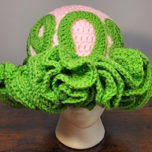 May include: A pink and green crocheted hat with a ruffled brim. The hat has a design of green circles on the pink crown.