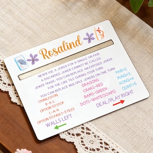 May include: A white game card with the name "Rosalind" in orange script, decorated with purple floral accents. The card includes game instructions and terms, with directional arrows and a slot.