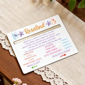 May include: A white game card with the name "Rosalind" in orange script, adorned with purple floral accents. The card features game instructions and a scoring guide, set against a neutral background.