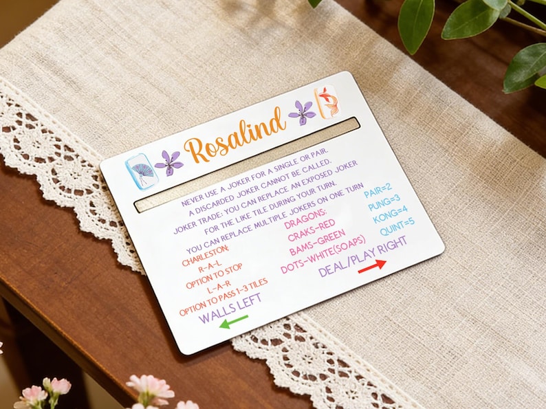 May include: A white game card with the name "Rosalind" in orange script, adorned with purple floral accents. The card features game instructions and a scoring guide, set against a neutral background.