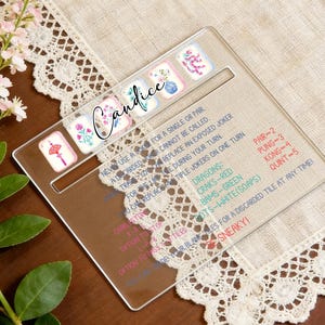 May include: A clear acrylic Mahjong tile holder with a personalized name, Candice, in cursive script. The holder features decorative tiles and game instructions. The background includes a floral arrangement and a lace-trimmed cloth.