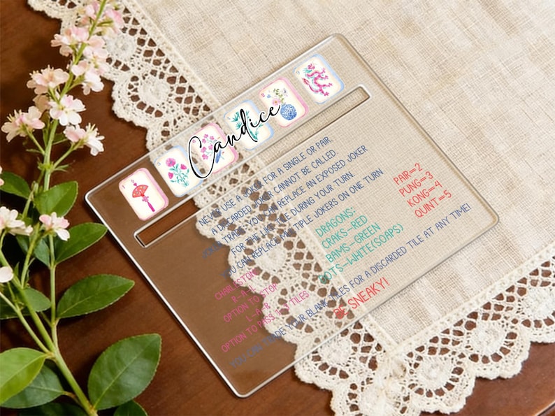 May include: A clear acrylic Mahjong tile holder with a personalized name, Candice, in cursive script. The holder features decorative tiles and game instructions. The background includes a floral arrangement and a lace-trimmed cloth.
