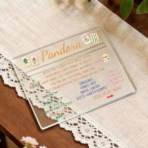 May include: A clear acrylic Mahjong game guide with the name "Pandora" in orange script. The guide includes rules, tile descriptions, and game instructions. It rests on a cream-colored cloth with a lace trim.