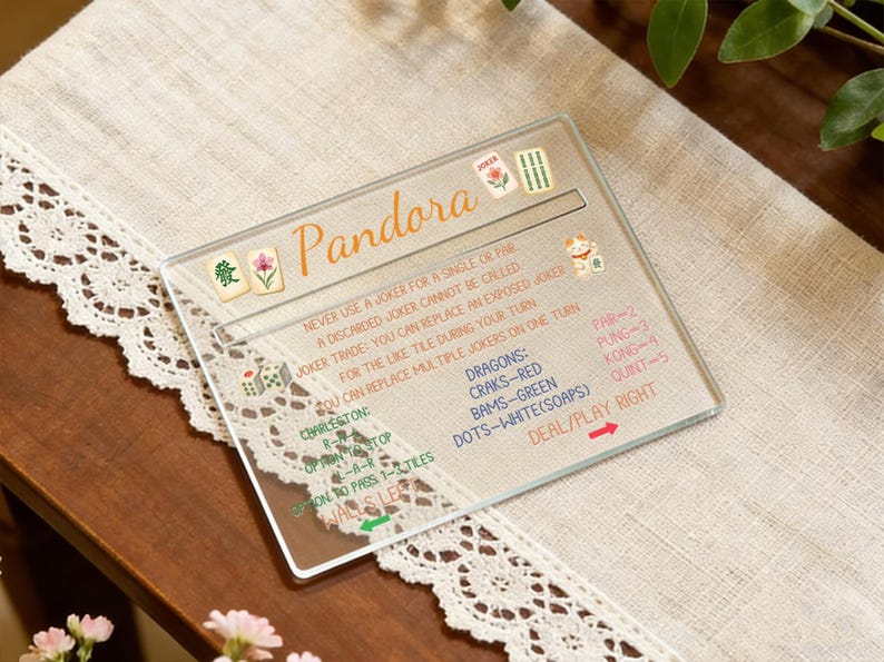 May include: A clear acrylic Mahjong game guide with the name "Pandora" in orange script. The guide includes rules, tile descriptions, and game instructions. It rests on a cream-colored cloth with a lace trim.