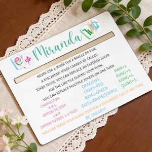 May include: A white Mahjong game card with the name "Miranda" in teal script. The card includes game rules and instructions, along with colorful tile illustrations. The card is on a cream-colored lace tablecloth.