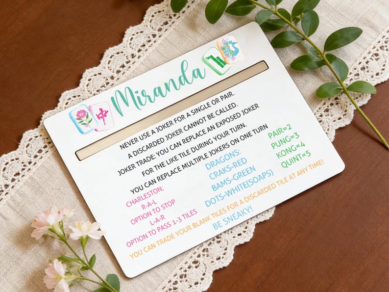 May include: A white Mahjong game card with the name "Miranda" in teal script. The card includes game rules and instructions, along with colorful tile illustrations. The card is on a cream-colored lace tablecloth.
