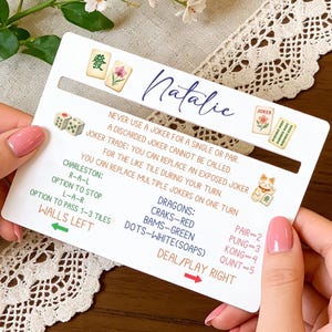 Puede incluir: Una tarjeta blanca con las reglas del juego de Mahjong y el nombre "Natalie" en cursiva. La tarjeta presenta ilustraciones de dados, fichas y un gato de la suerte. El texto incluye instrucciones del juego y detalles de puntuación.