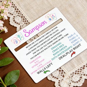 May include: A white game card with the name "Sampson" in pink script, featuring game instructions and floral illustrations. The card includes text about jokers, Charleston, and scoring. A decorative floral arrangement and lace trim surround the card.