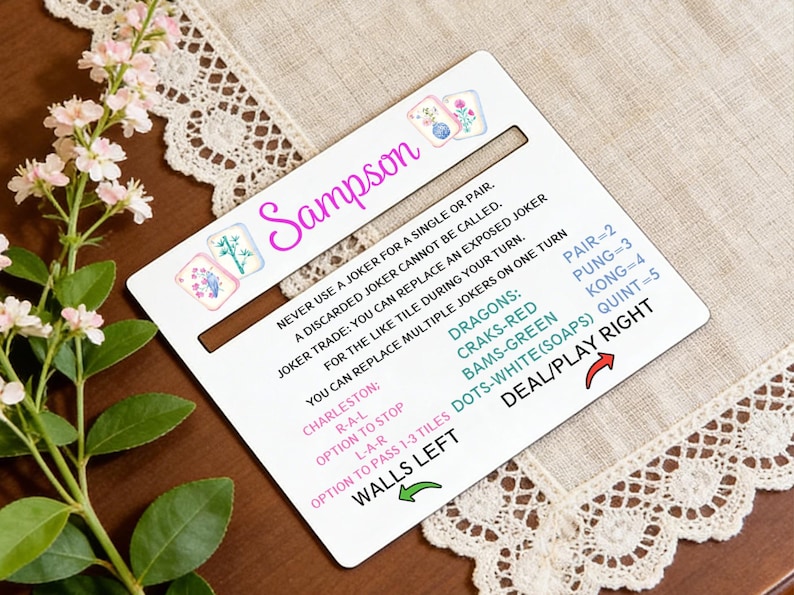 May include: A white game card with the name "Sampson" in pink script, featuring game instructions and floral illustrations. The card includes text about jokers, Charleston, and scoring. A decorative floral arrangement and lace trim surround the card.