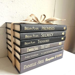 May include: A stack of eight black and gray hardcover books with gold lettering. The titles are "Promises", "Secrecy", "Treasures", "The Gift", "a painted house", and "Bungalow 2". The authors are Belva Plain and Danielle Steel.