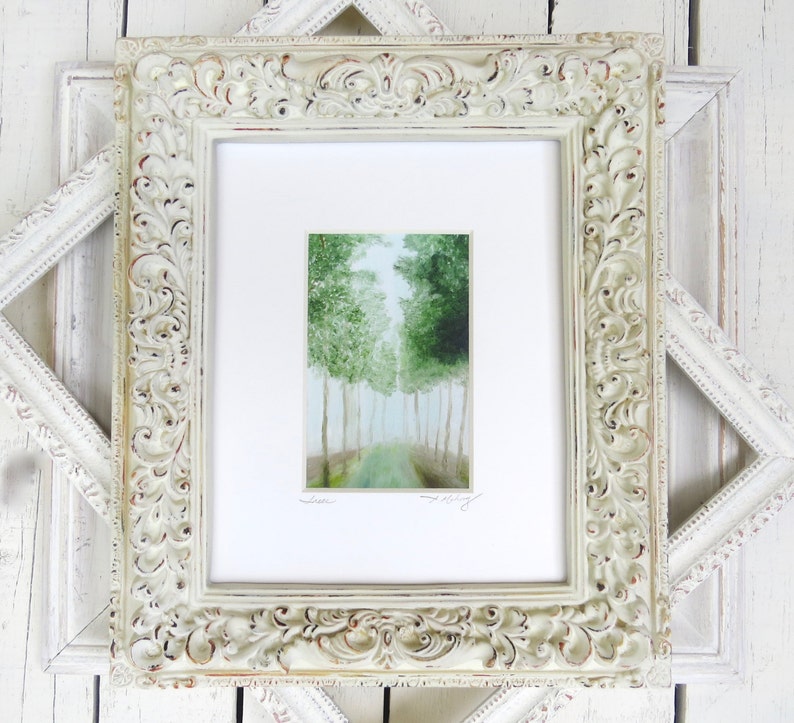 May include: A framed watercolor painting of a forest scene. The artwork features a path leading into a grove of green trees. The painting is set within a white mat and an ornate, off-white, distressed wooden frame.