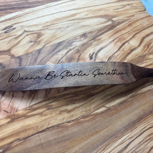 May include: Wooden spatula with the engraved text "Wanna Be Startin' Somethin" on a wooden cutting board. The spatula is dark brown with a handle and a hole for hanging. The cutting board has a light brown wood grain.