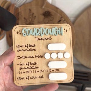May include: A square wooden sourdough timesheet with light blue 3D text and white markers. It features labels for Start of bulk fermentation, Stretch and Folds, End of bulk fermentation, and Start of cold rest, including temperature and time guides.
