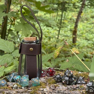 Könnte beinhalten: Eine transparente Glasflasche mit Korken, umhüllt von dunkelbraunem Leder und einem Griff. Verschiedene Würfel in Grün, Rot und Schwarz deuten auf ein Tabletop-Gaming-Thema hin. Die Flasche ist etwa 15 cm hoch.