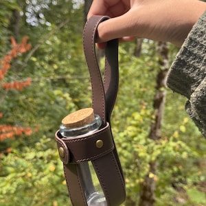 May include: A clear glass bottle with a cork stopper, held in a brown leather carrier with a handle. The carrier has brass-coloured rivets. The background is a blurred view of a forest with green foliage.