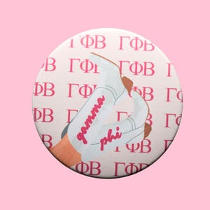 May include: A round button with a pink background and a white circle in the center. The circle features a repeating pattern of Greek letters and a pair of white cowboy boots with the words "gamma phi" in pink script.